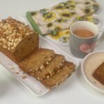 A freshly baked applesauce bread topped with oats and walnuts, sliced and served on a white platter. A cup of tea, a sunflower-patterned oven mitt, and a small vase with pink flowers complete the cozy setup.