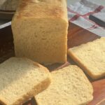Loaf of homemade white sandwich bread on a wooden cutting board, with three even slices fanned out in soft sunlight; a striped kitchen towel and glass cloche sit in the background.