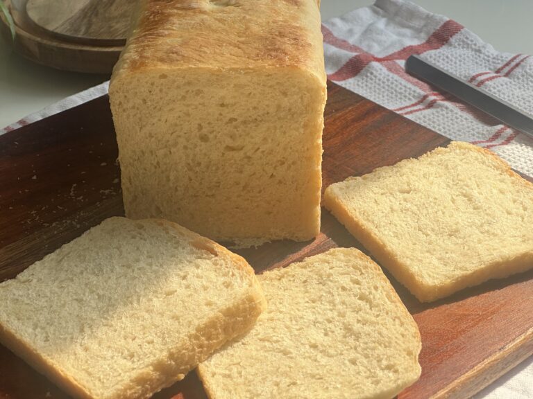 🍞 How to Make the Perfect White Bread at Home – Soft, Fluffy & Bakery-Style Loaf of homemade white sandwich bread on a wooden cutting board, with three even slices fanned out in soft sunlight; a striped kitchen towel and glass cloche sit in the background.