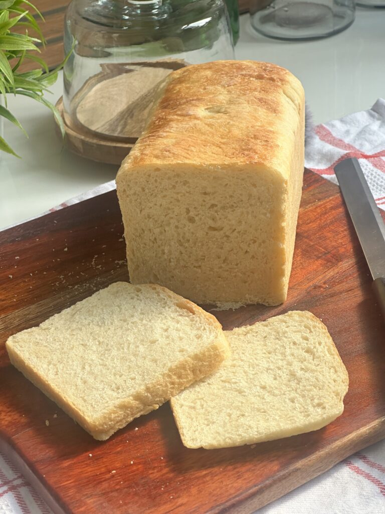 “Freshly baked white sandwich loaf on a wooden cutting board, with two even slices fanned out in front, a bread knife alongside, and a kitchen towel and glass cloche in the softly lit background.”