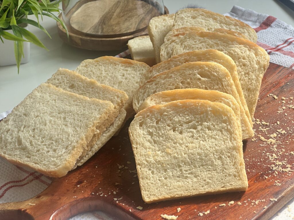Multiple even slices of homemade white sandwich bread fanned out on a wooden cutting board, with golden crumbs scattered around; a striped tea towel, glass cloche, and potted plant sit softly blurred in the background.