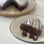 Close-up of a rich, moist French chocolate cake (Gâteau au Chocolat) topped with fresh raspberries and powdered sugar, placed on a white plate.