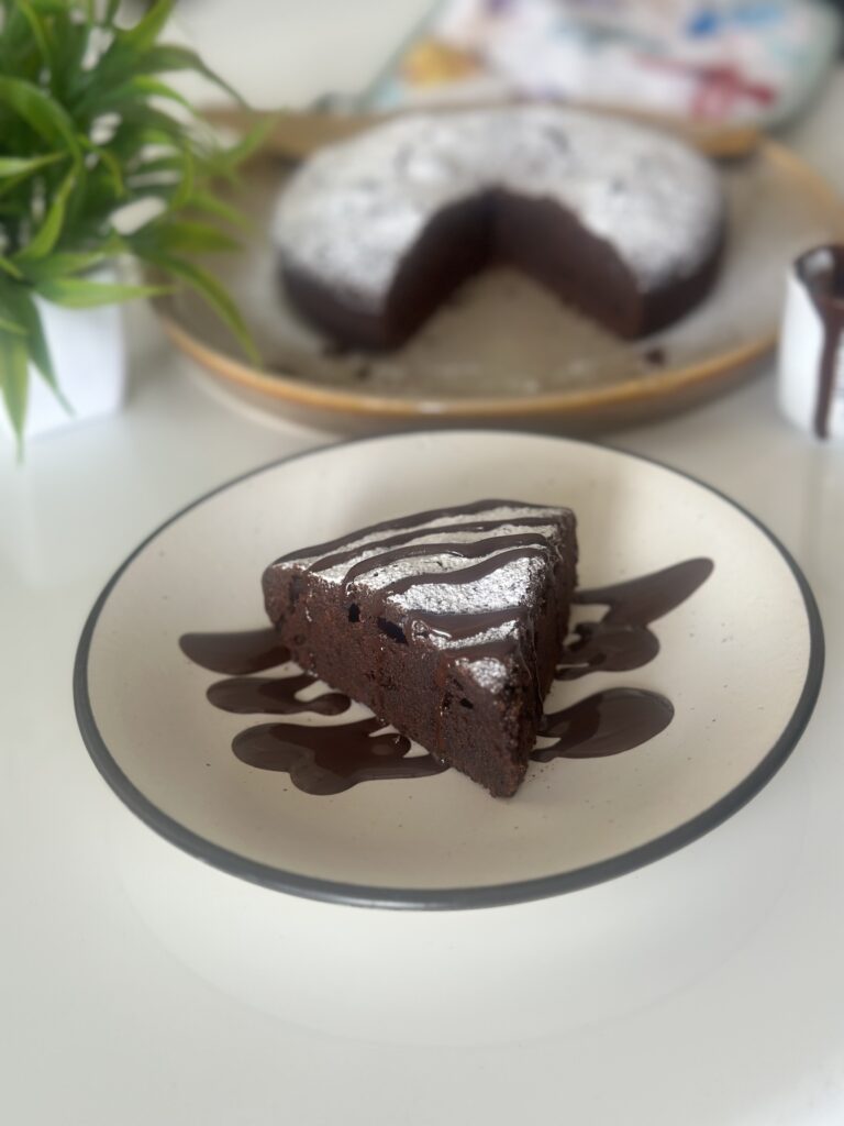 A slice of rich French Gâteau au Chocolat drizzled with chocolate sauce and dusted with powdered sugar, served on a plate with the full cake in the background.