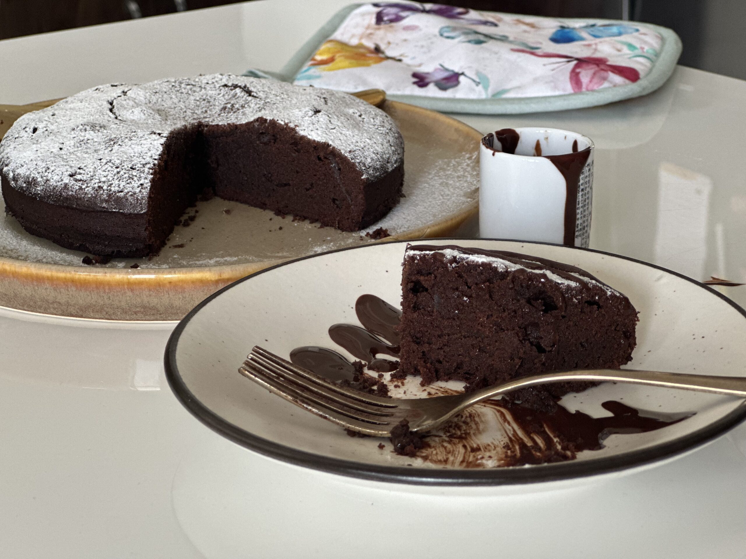 A slice of rich French chocolate cake served with chocolate sauce, with the whole gâteau au chocolat dusted with powdered sugar in the background.