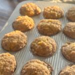 Freshly baked mango muffins in a silver muffin tray, golden and sugar-crusted, glowing warmly in the sunlight with a sunflower-patterned cloth in the background.