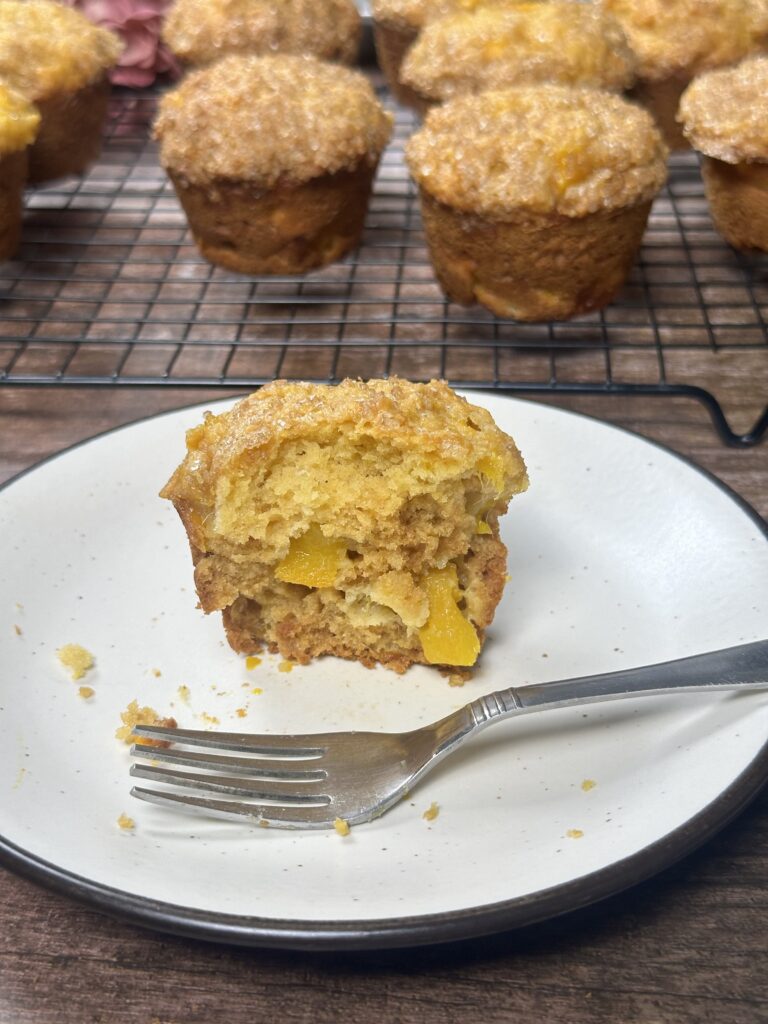 Half-eaten mango muffin with egg, showing soft crumb and juicy mango pieces on a white plate with a fork, surrounded by whole muffins cooling on a wire rack.