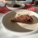 A single slice of chocolate brownie on a cream ceramic plate, with sunlight casting soft shadows and additional brownie pieces blurred in the background.