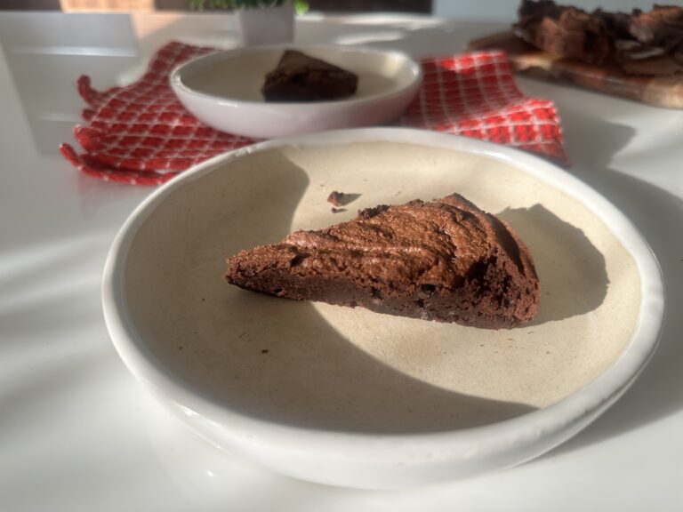 Easy Air Fryer Brownies — Fudgy, Gooey & Ready in 15 Minutes! A single slice of chocolate brownie on a cream ceramic plate, with sunlight casting soft shadows and additional brownie pieces blurred in the background.