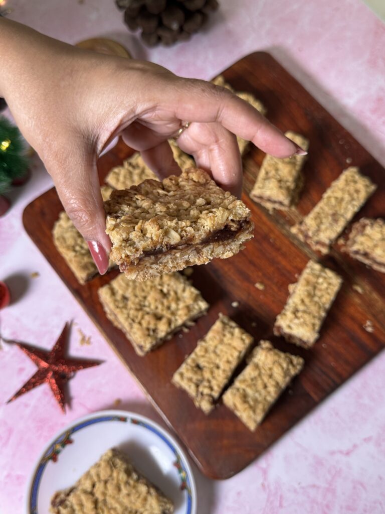 Soft & Buttery Jam Bars – A Simple Christmas Treat for Bakemas 2025 Hand holding a soft and buttery strawberry jam bar, showing layers of crumbly oat topping, fruity jam filling, and tender base, with more jam bars arranged on a wooden board in the background.