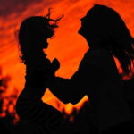 Silhouette of a mother lifting her young daughter against a glowing red sunset sky, expressing love and connection.