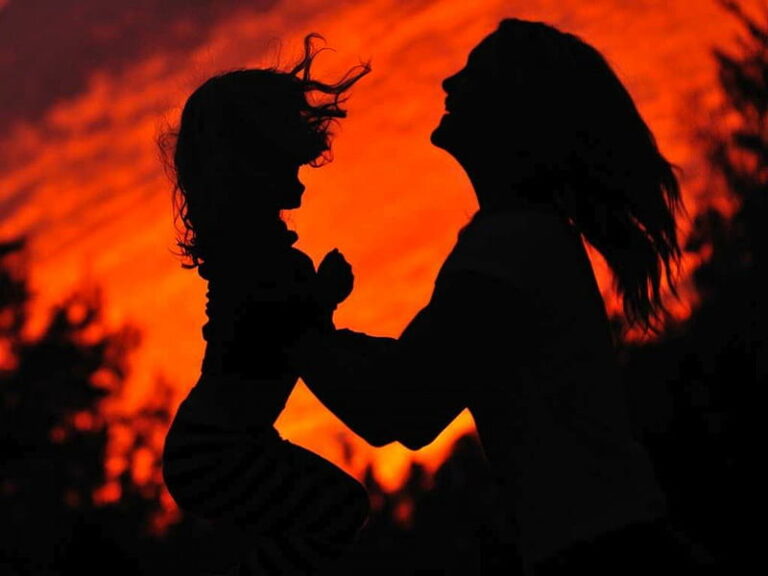 Silhouette of a mother lifting her young daughter against a glowing red sunset sky, expressing love and connection.