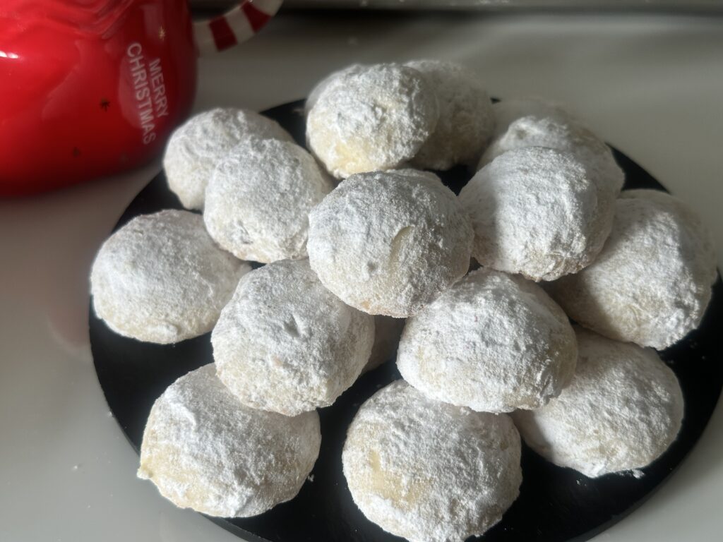 Overhead view of classic melting moments cookies coated in powdered sugar, arranged on a black plate and styled with a festive Christmas wreath and a red holiday mug.