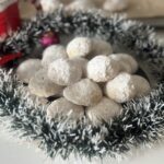 A plate of powdered sugar–coated melting moments cookies arranged like snowballs, surrounded by a silver Christmas wreath with soft natural light.