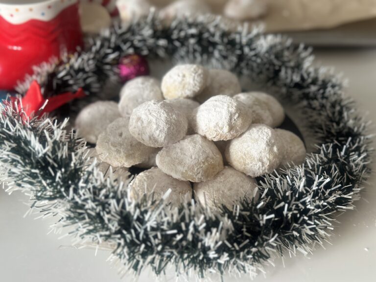 A plate of powdered sugar–coated melting moments cookies arranged like snowballs, surrounded by a silver Christmas wreath with soft natural light.