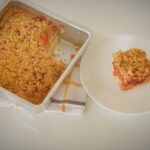 Strawberry streusel cake in a square baking pan with a slice served on a white plate, showing layers of soft cake, strawberry filling, and crumb topping.