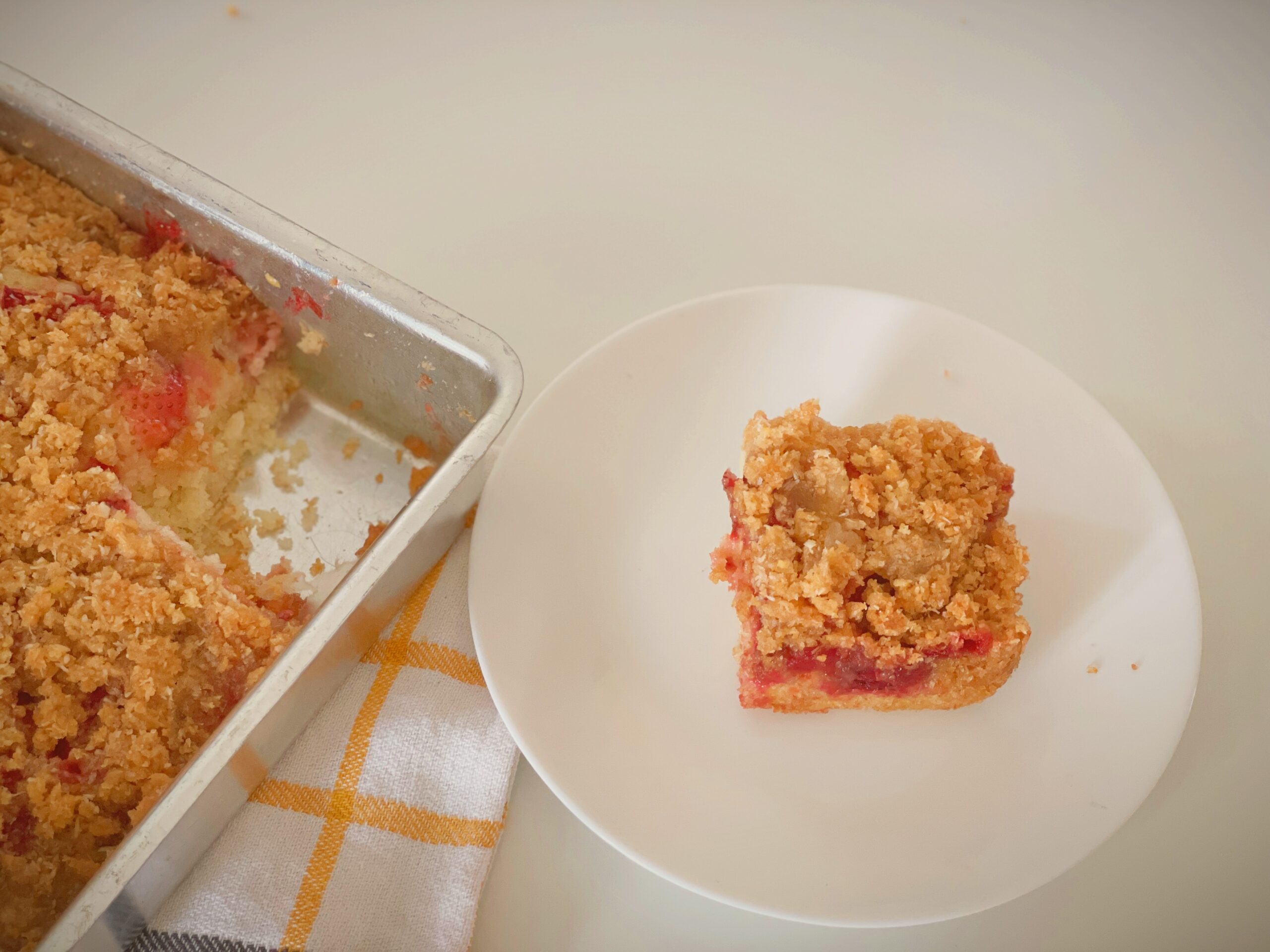 Slice of strawberry streusel cake on a white plate with remaining cake in a baking pan, showing strawberry layer and crumb topping.