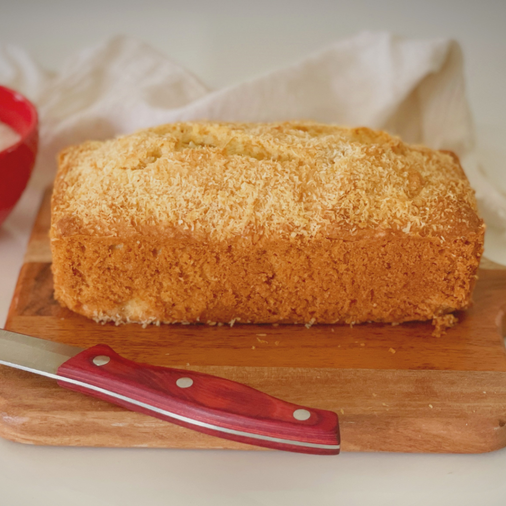 Golden coconut loaf cake on a wooden board with a knife in front, topped with desiccated coconut and showing a soft, moist crumb.