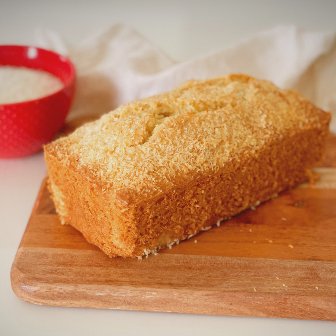 Golden coconut loaf cake topped with desiccated coconut placed on a wooden board, with a blurred bowl of ingredients in the background.