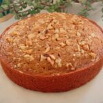 Eggless coffee walnut cake on a white plate with toasted walnuts on top, surrounded by green leaves