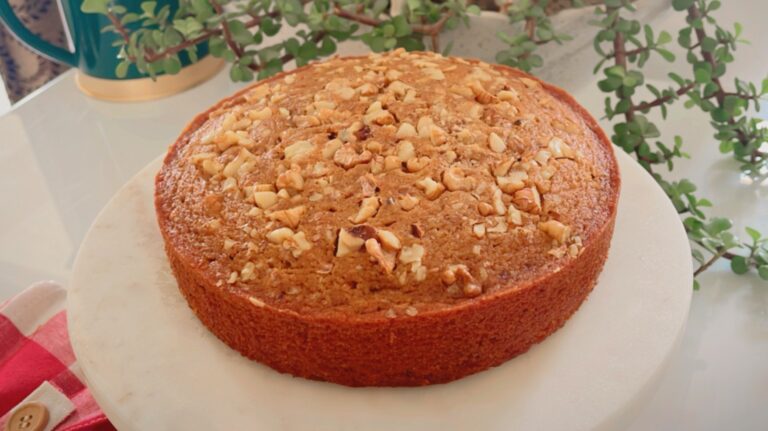 Eggless coffee walnut cake on a white plate with toasted walnuts on top, surrounded by green leaves