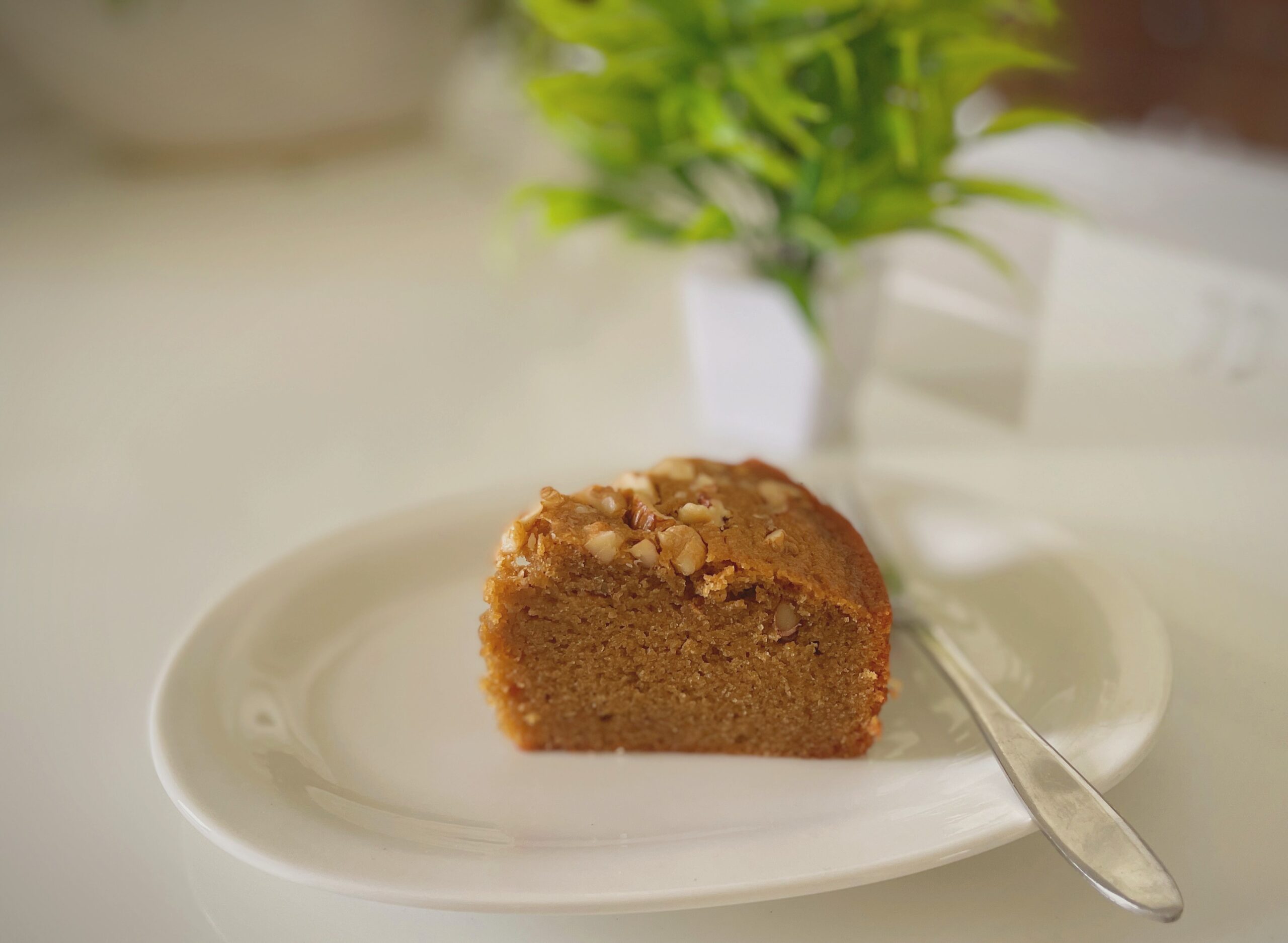 A slice of eggless coffee walnut cake on a white plate with a fork beside it and a green plant softly blurred in the background