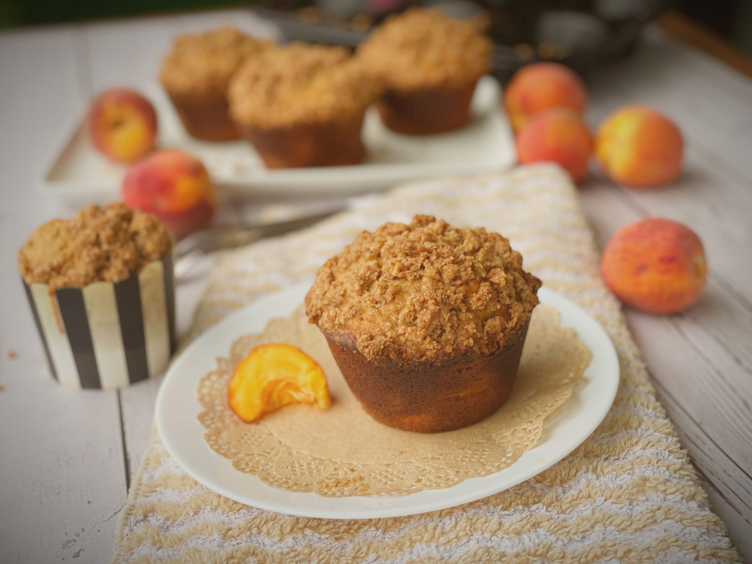Brown butter peach cobbler muffin plated on a white decorative doily with fresh peach slice garnish and striped paper liner visible