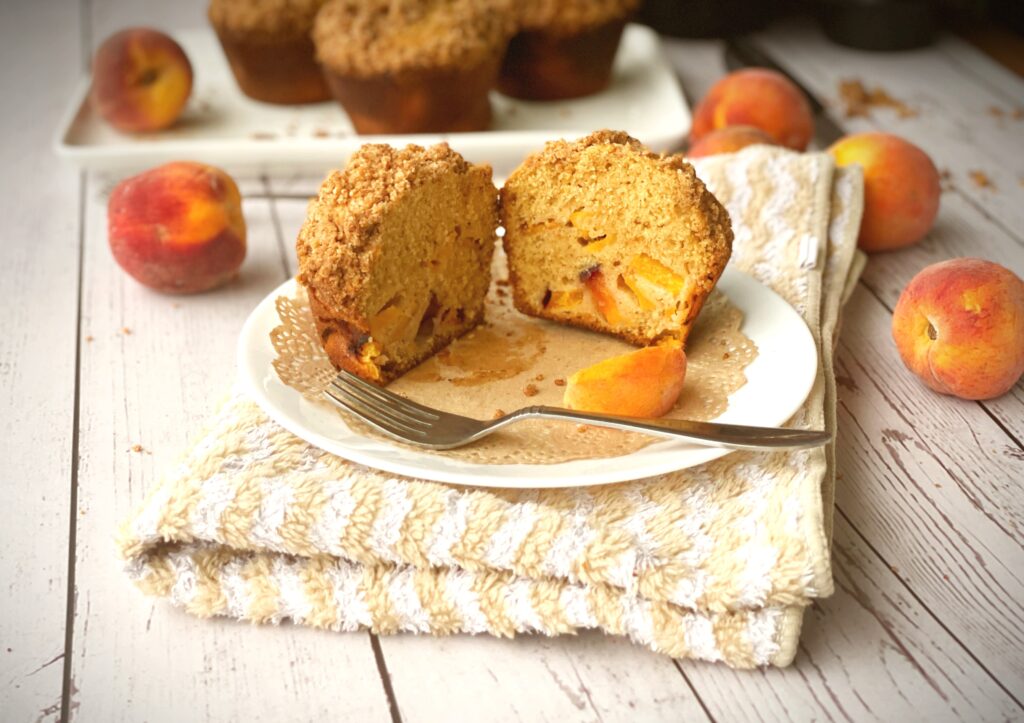 Brown butter peach cobbler muffin cut in half showing fluffy interior with peach pieces, served on a white plate with fork and fresh peach garnish