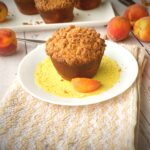 Brown butter peach cobbler muffin with golden streusel topping on a yellow doily with fresh peaches