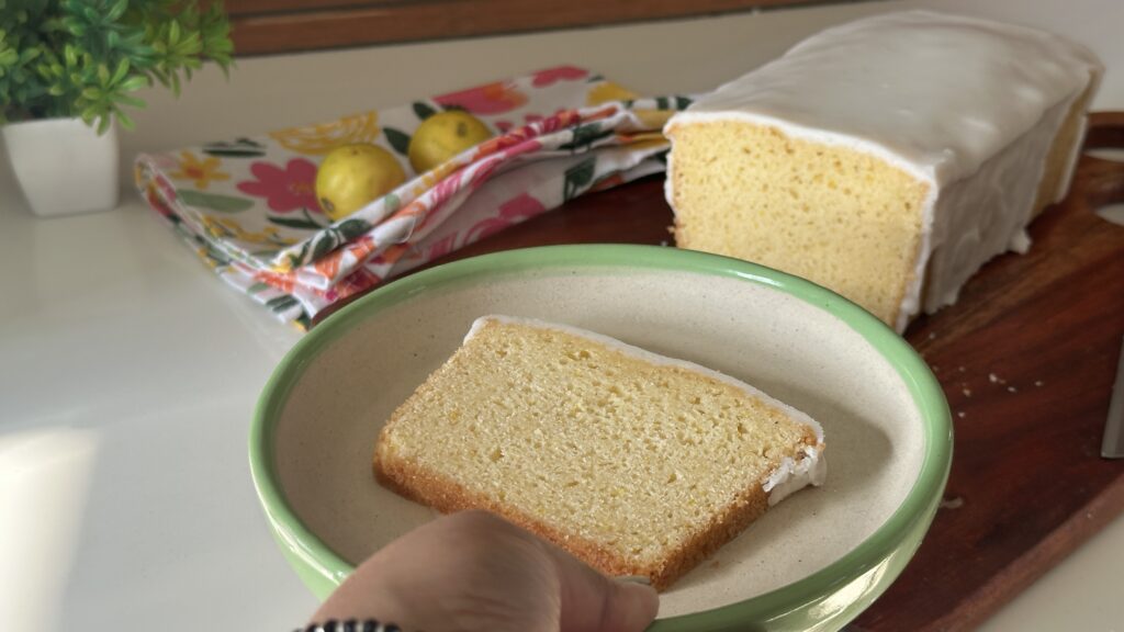 Slice of Mary Berry lemon drizzle cake in bowl with glazed loaf in background and fresh lemons