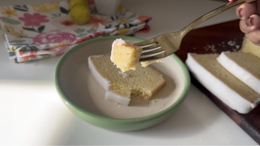 Fork holding a bite of Mary Berry lemon drizzle cake with icing glaze over sliced cake in bowl