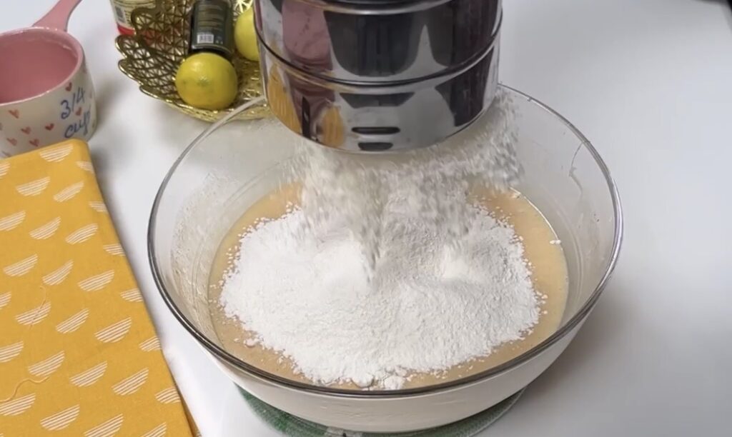 Flour being sifted into condensed milk cake batter in a glass bowl