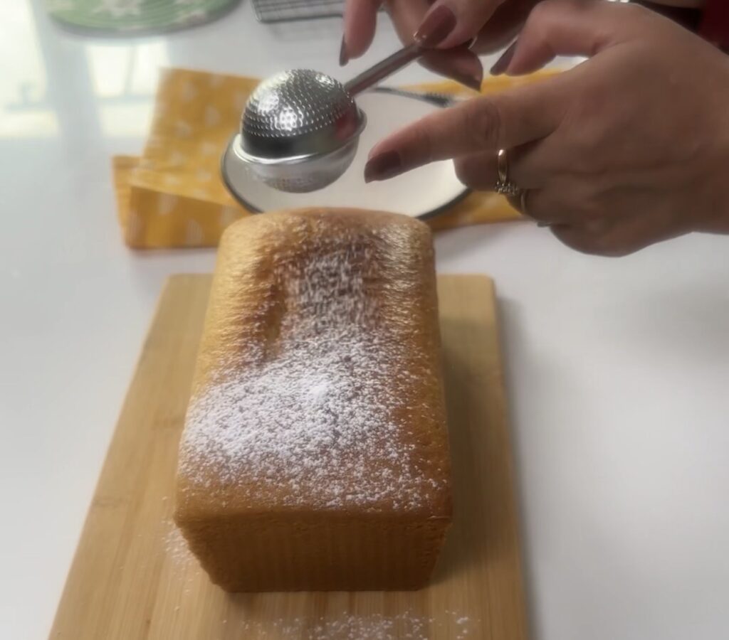 Powdered sugar being dusted over a golden condensed milk loaf cake on a wooden board