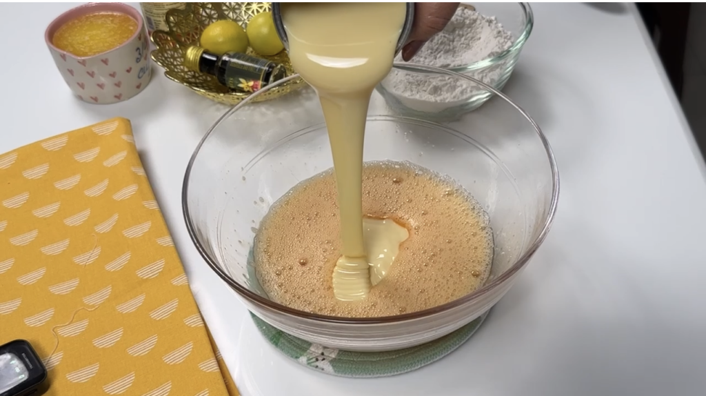 Sweetened condensed milk being poured into whisked eggs in a glass bowl for condensed milk cake