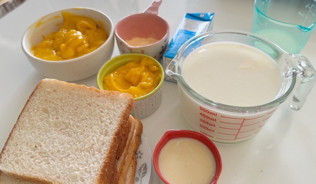 Ingredients for eggless mango bread pudding arranged on a table, including white bread slices, fresh mango puree, milk, condensed milk, cream, and custard powder.
