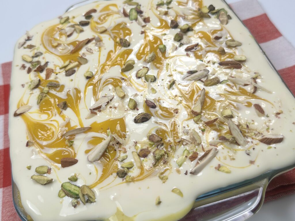 Close-up of creamy eggless mango bread pudding topped with mango swirls, chopped pistachios, and almond slivers in a glass dish.