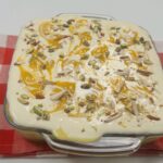 Creamy eggless mango bread pudding in a square glass dish, topped with swirls of mango puree, chopped pistachios, and almond slivers, served as a chilled no-oven summer dessert.