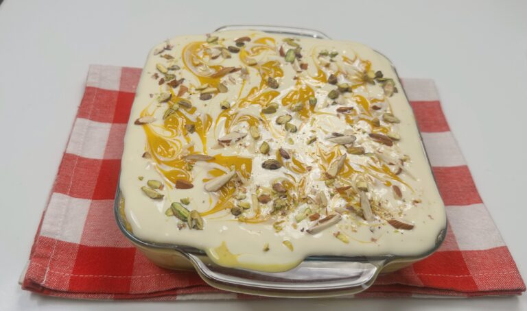 Creamy eggless mango bread pudding in a square glass dish, topped with swirls of mango puree, chopped pistachios, and almond slivers, served as a chilled no-oven summer dessert.