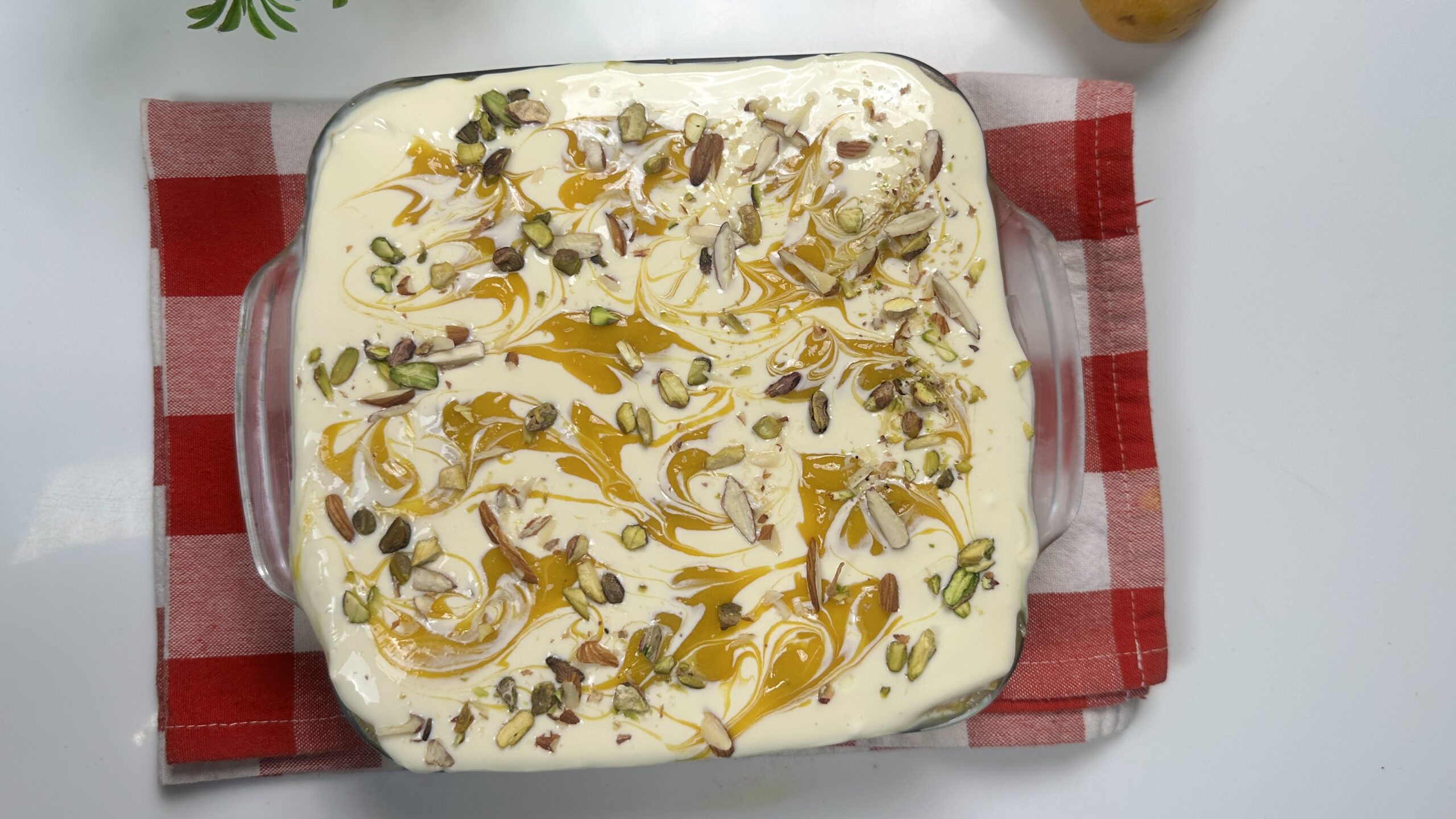 Top view of creamy eggless mango bread pudding in a glass baking dish, decorated with mango swirls, chopped pistachios, and almond slivers on a red checkered cloth.