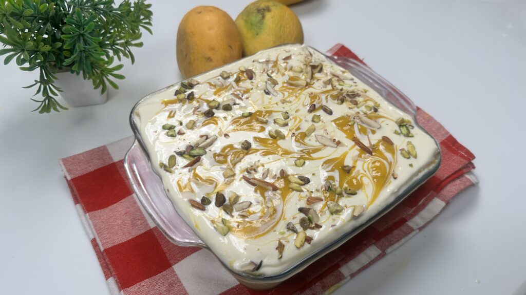 Eggless mango bread pudding in a glass dish topped with mango swirls, pistachios, and almond slivers, placed on a red checkered cloth with fresh mangoes in the background.