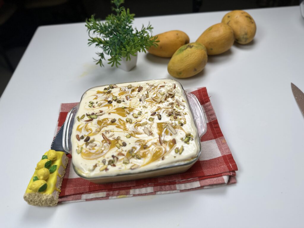 Eggless mango bread pudding in a glass dish topped with mango swirls, pistachios, and almond slivers, placed on a red checkered cloth with fresh mangoes in the background.