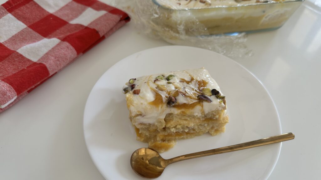 A creamy slice of mango bread pudding served on a white plate with a golden spoon, topped with mango swirls and chopped pistachios, with the pudding dish blurred in the background.