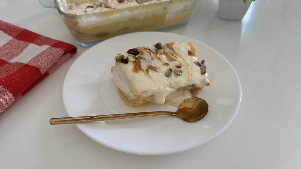 A chilled serving of eggless mango bread pudding on a white plate with a golden spoon, topped with mango swirls and chopped pistachios, with the dessert dish blurred in the background.
