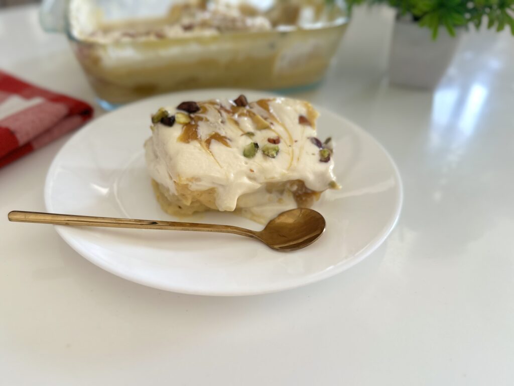 A creamy slice of eggless mango bread pudding served on a white plate with a golden spoon, topped with mango swirls and chopped pistachios.