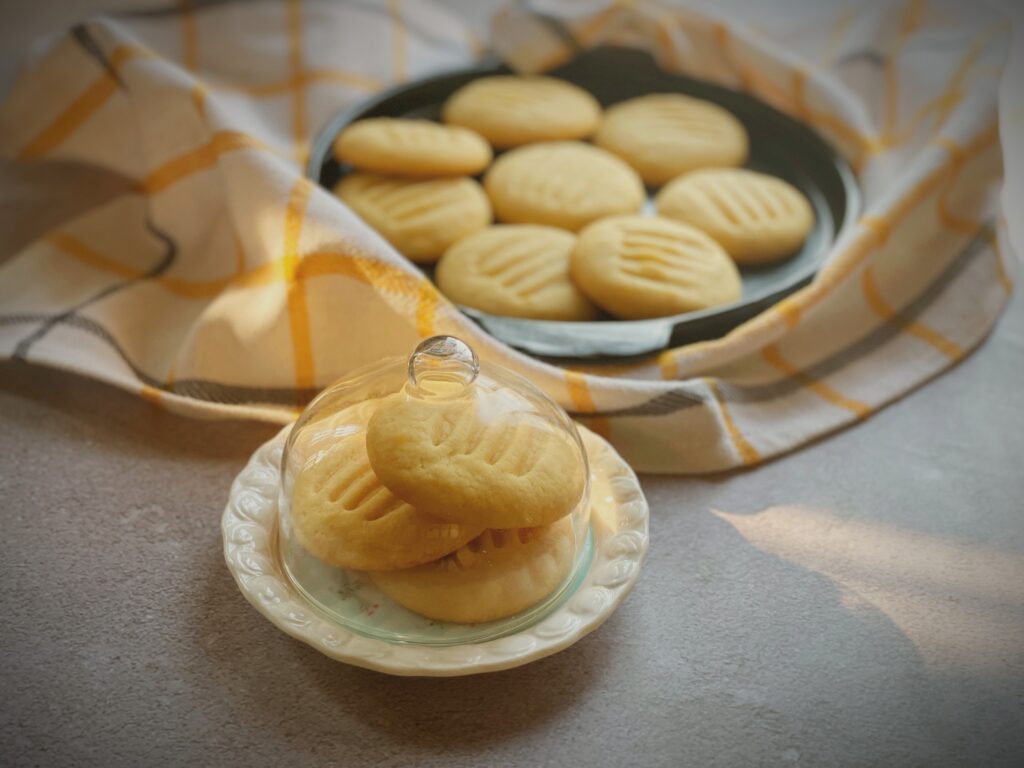 Eggless condensed milk cookies displayed in a small glass butter dish dome beside a platter on a yellow checkered cloth