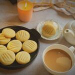 Eggless condensed milk cookies on dark plate beside a cup of chai, white teapot, butter dish and lit candle