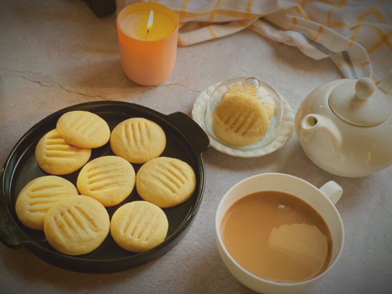 Eggless condensed milk cookies on dark plate beside a cup of chai, white teapot, butter dish and lit candle