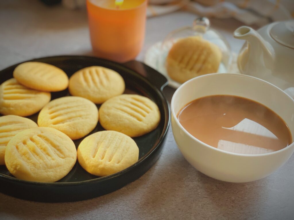 Eggless condensed milk cookies served on dark plate beside a steaming cup of chai, white teapot and candle