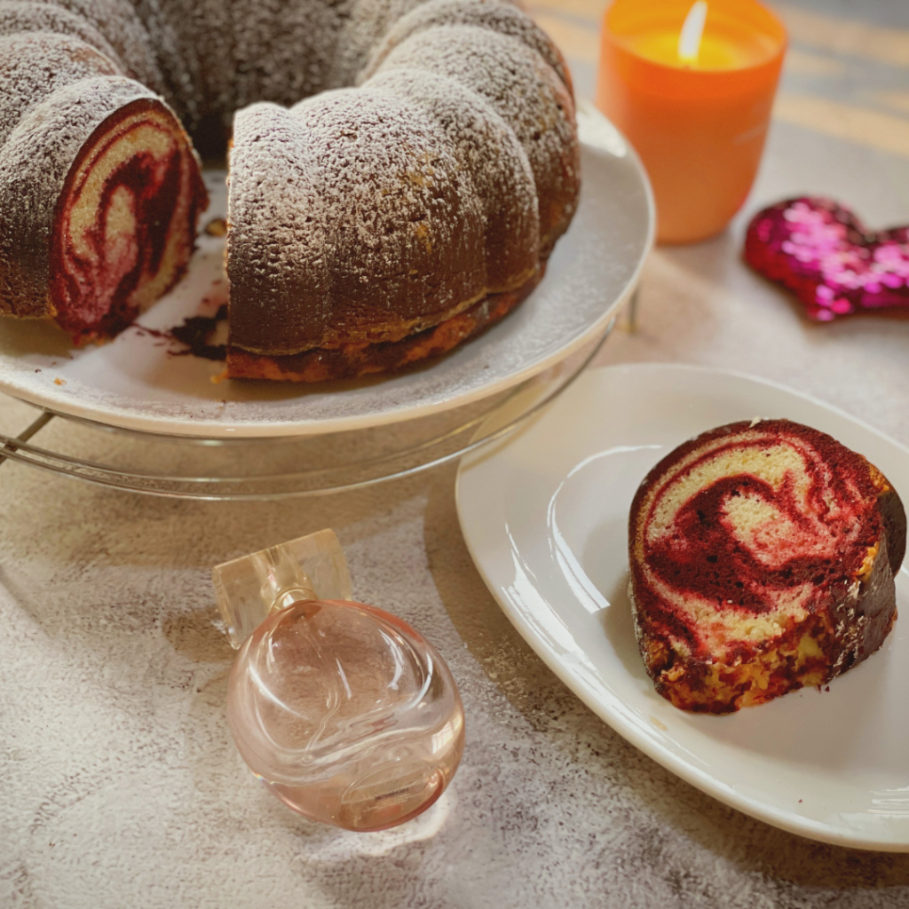 Red velvet marble cake on a stand next to a single slice on a plate, with a lit candle and perfume bottle in the background