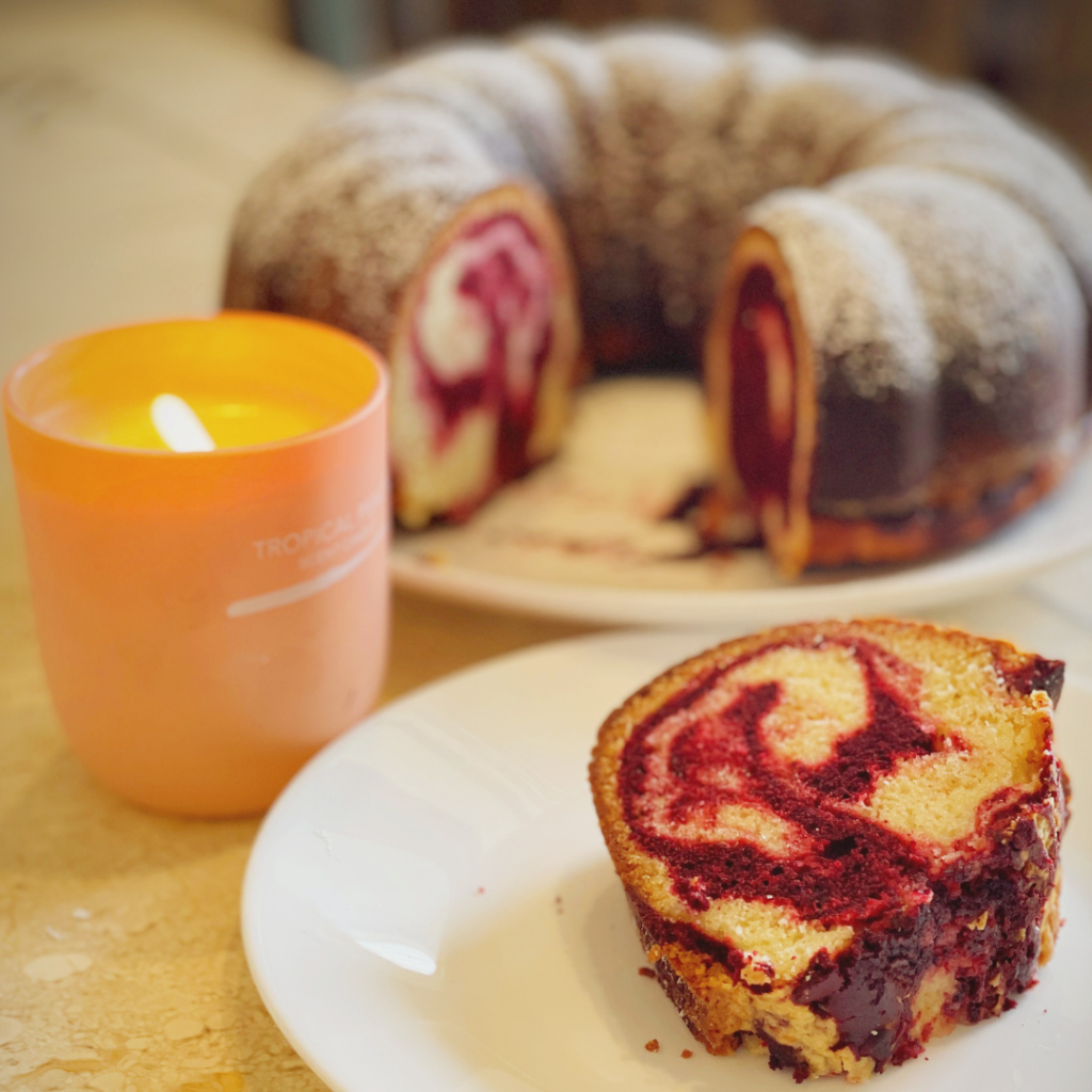 Close-up of a red velvet marble cake slice on a plate beside a lit candle, with the whole cake blurred in the background