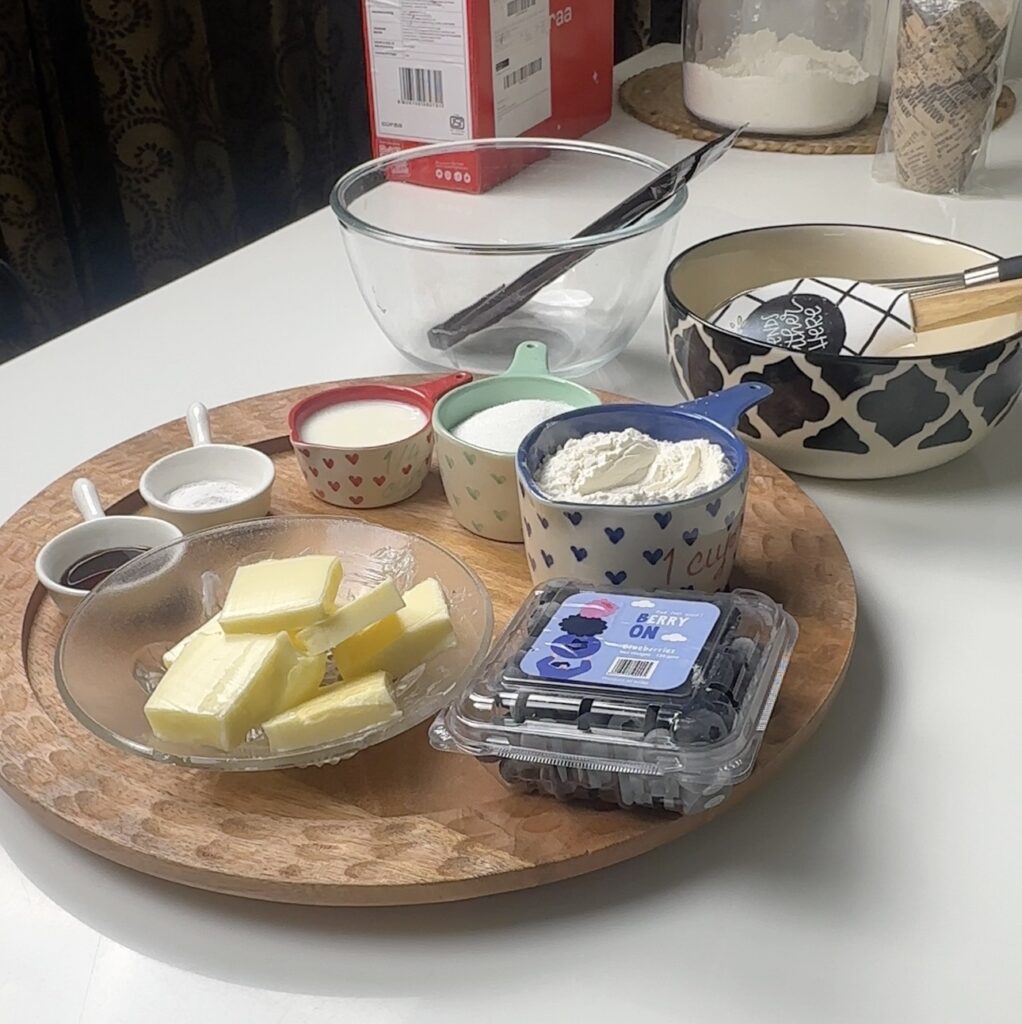 Ingredients for Blueberry Chocolate Muffins laid out on a wooden tray — butter, flour, sugar, milk, blueberries